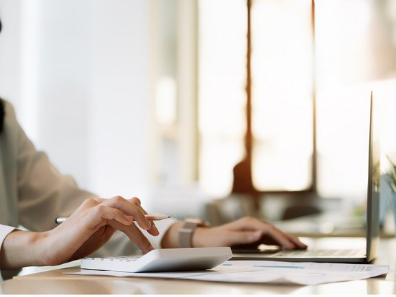 Nahaufnahme einer Geschäftsfrau, die im modernen Büro Berechnungen mit Taschenrechner und Laptop durchführt.